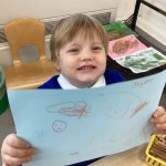 a child is proudly holding up a drawing they have created on a blue sheet of paper, with a name "Taylor" written on it.