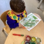 a child wearing a bib is seated at a table, focused on painting a picture with green and yellow watercolors.