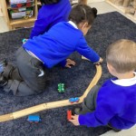 two children are on the floor, working on connecting a wooden train track together and playing with toy trains.
