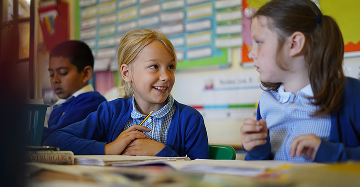 Three Dartford Primary Academy students are shown sat at a desk. Two of them are girls who are looking towards one another with smiles.