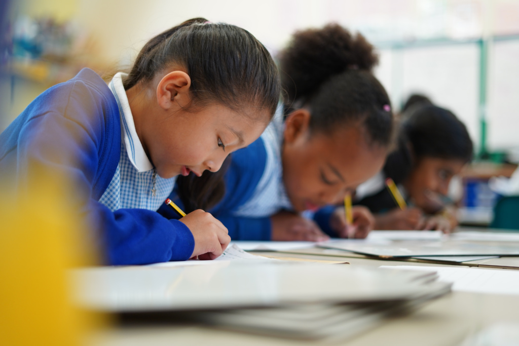 Three female Dartford Primary Academy students are shown sat at a desk and writing on paper with pencils.