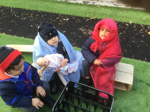 Three Nursery pupils are pictured sat outdoors wearing blankets to act out the Nativity story with toy baby doll.