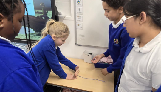 Three female pupils are pictured gathered round a desk, listening to one of their peers who is seen demonstrating something to them on World Maths Day.