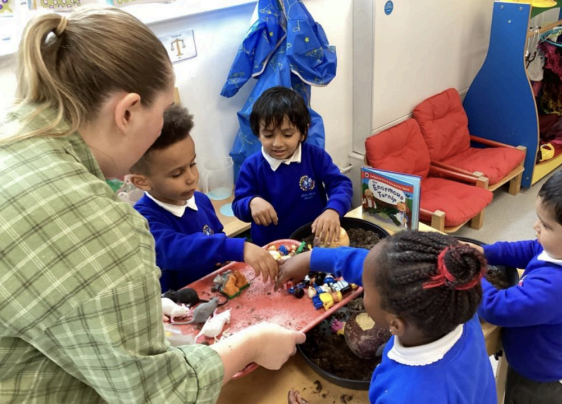 A group of four Nursery pupils are pictured sitting down together, alongside their class teacher, playing with some toys.