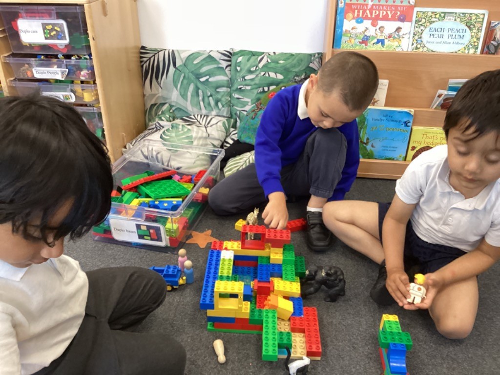 In the image, three young boys are sitting on a carpeted floor, engrossed in playing with a large pile of colorful building blocks, likely LEGO or Duplo. They have a clear plastic container filled with more blocks next to them. One boy, on the left, is wearing a white polo shirt and is actively building with the blocks. The other boy, on the right, is wearing a blue long-sleeved top and dark trousers, and is also engaged in building, with some animal figures also visible among the blocks.