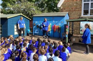 This image depicts an outdoor educational or entertainment activity, likely at a school. A large group of young children, predominantly wearing blue tops, are sitting on the ground, facing a small raised wooden stage or platform. On the platform, two adults are present. One adult, wearing a blue t-shirt and black trousers, is standing on the left side of the platform. The other adult, also in a blue top, is standing more centrally and appears to be leading the activity, possibly interacting with the children. Behind the stage, there are two blue wooden sheds. The shed on the right has various musical instruments or sound-making objects attached to its exterior, such as pans, cymbals, and colorful tubes, suggesting it might be a "sound wall" or similar interactive display. To the far right, another adult, dressed in a blue jacket, is standing near a wall or building. The setting appears to be a school playground, with brick buildings visible in the background and trees overhead.