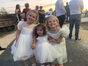 Three young children in white dresses are standing close together and holding hands outdoors. There are adults and a vehicle in the background, suggesting some kind of gathering