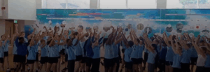 A large group of children, likely students, are gathered indoors, possibly in a school hall or gymnasium. They are all raising their arms above their heads. There's a large mural or display on the wall behind them that appears to be a timeline.