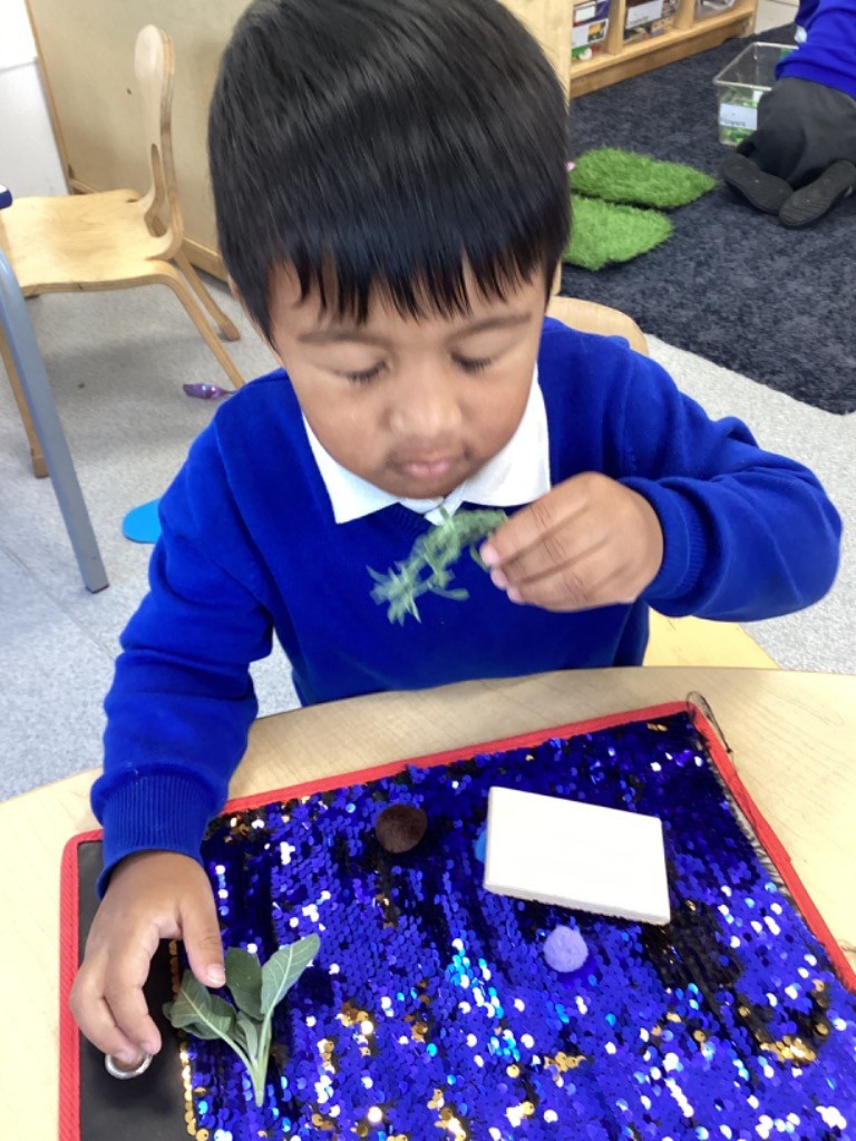 A boy is handling a sprig of green leaves/herbs and a small, spiky plastic object (possibly a toy insect or plant) over a reversible sequin mat (blue and gold), engaging with various textures and objects.