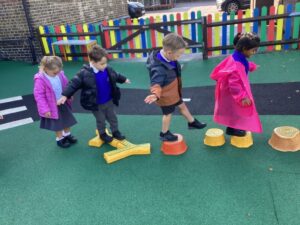 A group of children is playing on an outdoor playground. They are walking and balancing on a series of brightly colored stepping stones and a wooden beam.