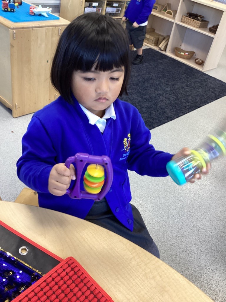 A girl is holding two different noise/sensory toys: a purple rattle/shaker and a clear bottle shaker filled with beads or small objects, engaging in sound-making and visual observation.