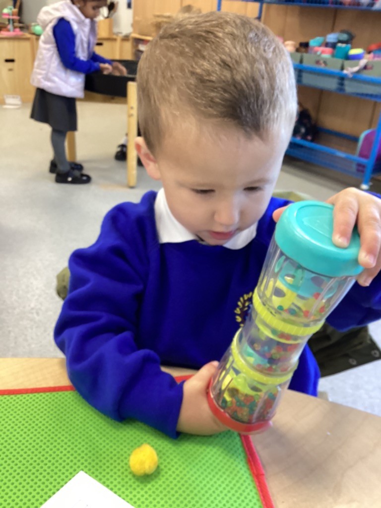 A boy is holding a clear, cylinder-shaped shaker toy that has various colorful elements inside. He is closely observing the movement and listening to the sound, another activity centered on sight and sound.