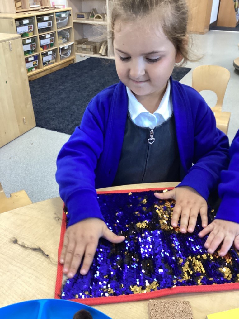 A girl is seated at a table, actively touching a rectangular mat covered in reversible sequins (blue and gold), which is a common item for tactile sensory play.