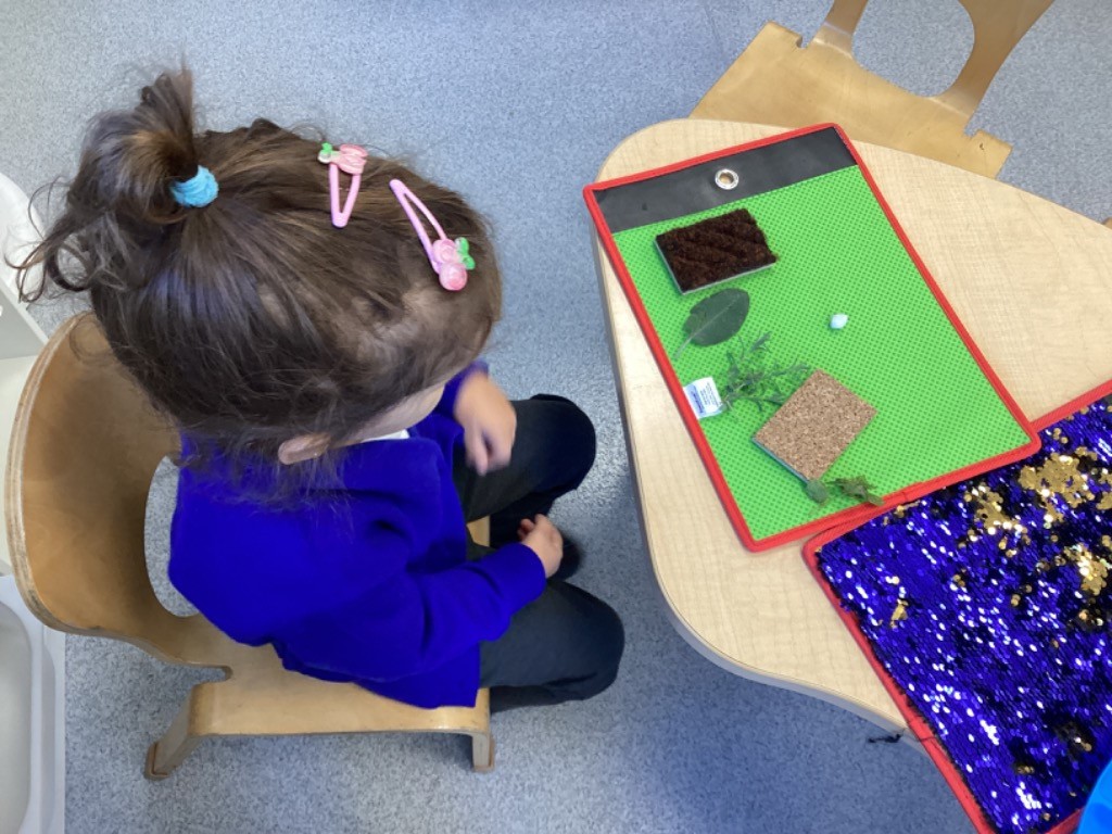 Taken from an overhead view, a girl is seated at a small table where a green mat is laid out with different sensory items, including a leaf, a small piece of cork, and some brown material (possibly soil or moss). This is another example of a sensory exploration station.