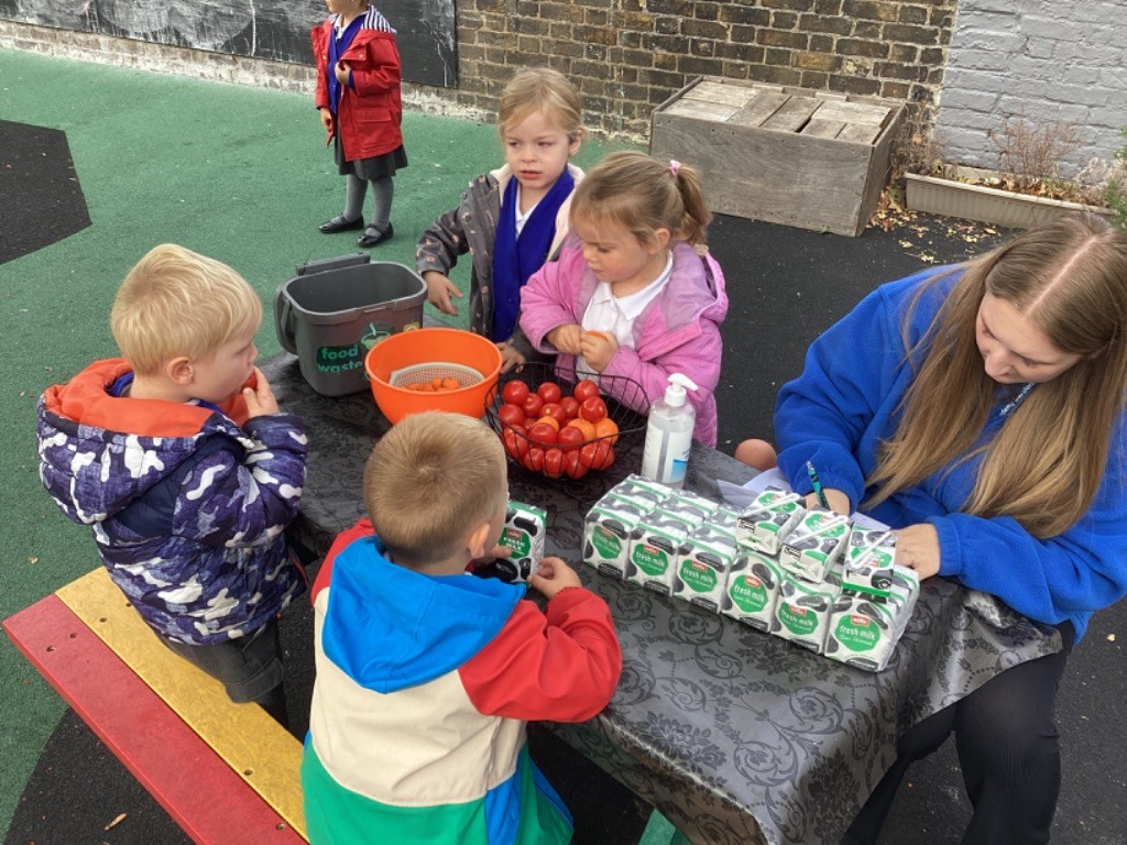 This image shows children and an adult outdoors around a table. They appear to be engaged in a food preparation or sorting activity, as there are buckets, a colander, and fresh tomatoes on the table. There are also multiple cartons of "fresh milk," suggesting they might be preparing for a snack or a lesson about food and nutrition.