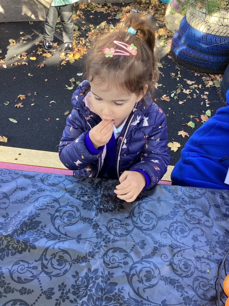 A girl in a puffy coat is outdoors, standing by a table with a dark patterned tablecloth, and is eating a small piece of food (possibly a piece of fruit or vegetable) from the items on the table,