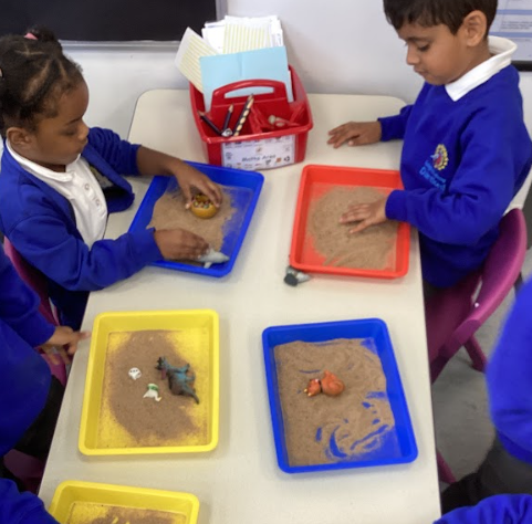 This image shows two young children at a table, each engaging in a hands-on learning activity. They are using trays filled with a substance that looks like sand, with small figurines of animals placed inside. This appears to be a sensory-based activity, possibly related to storytelling, creative play, or learning about animals, where the children can explore and interact with the materials.