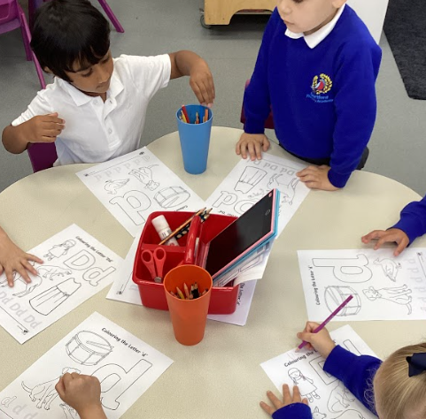 This image shows a group of children sitting at a round table, engaged in a coloring and writing activity. They each have a worksheet with large outline letters to color, specifically the letter "p." They have a pot of colored pencils in the center of the table. This is a fine motor skill and literacy-building activity, where the children are practicing letter recognition and coloring.