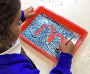 This image shows a young child using a brush to trace a large letter "m" in a red tray filled with blue and gold glitter. This is a sensory activity designed to help children learn to form letters in a creative, tactile way. The child is using a brush to carefully draw the letter in the glitter.