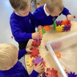 three children are seated around a table and are collaborating to build something with colorful interlocking gears.