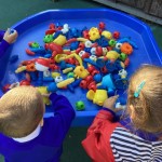a group of children are gathered around a blue tray, playing with a variety of colorful building blocks and toys.