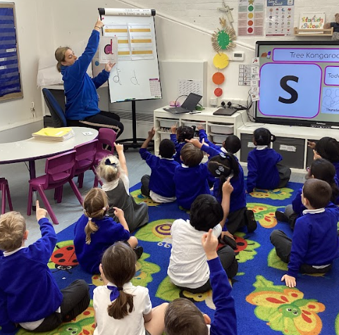 This image shows a classroom setting where a teacher is leading a group of young students. The teacher is standing in front of a whiteboard and pointing to the letter "d," while the children are sitting on a patterned rug, actively participating by raising their hands. A large screen to the right also displays a letter, "S." This appears to be a phonics lesson where the teacher is introducing or reviewing letters and sounds.
