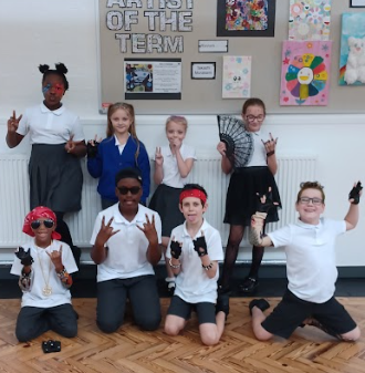 This group is clearly embracing a rock-and-roll aesthetic. Several children are wearing sunglasses, and some have temporary tattoo sleeves or arm covers. Accessories like bandanas, fingerless gloves, chains or necklaces, and brightly colored hair clips or wigs are visible. Two of the boys kneeling in the front row are dressed in a hip-hop/rock fusion style with bandanas, shades, and thick necklaces. One child is dressed in a style reminiscent of Michael Jackson (dark wig, sunglasses, black and white outfit). The overall mood is playful and cool, with the children posing confidently.