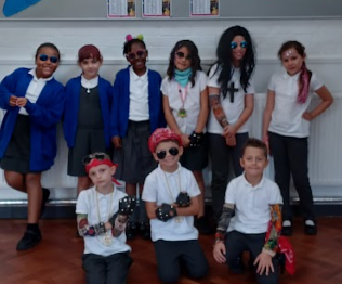 This group seems to be continuing the music/rock star theme, with many children making the "rock on" hand gesture (devil horns). The children are primarily wearing their school uniforms (white shirts, dark skirts or trousers) but have added themed accessories. Fingerless gloves are a common accessory among the children. Other accessories include bandanas, sunglasses, and a fan (held by the girl on the far right). Some children have face paint or makeup, such as the girl on the far left with red and blue paint. They are posing energetically, adding to the rock star vibe. The poster on the wall mentions "ARTIST OF THE TERM", which might suggest the day is linked to an arts or music appreciation event.