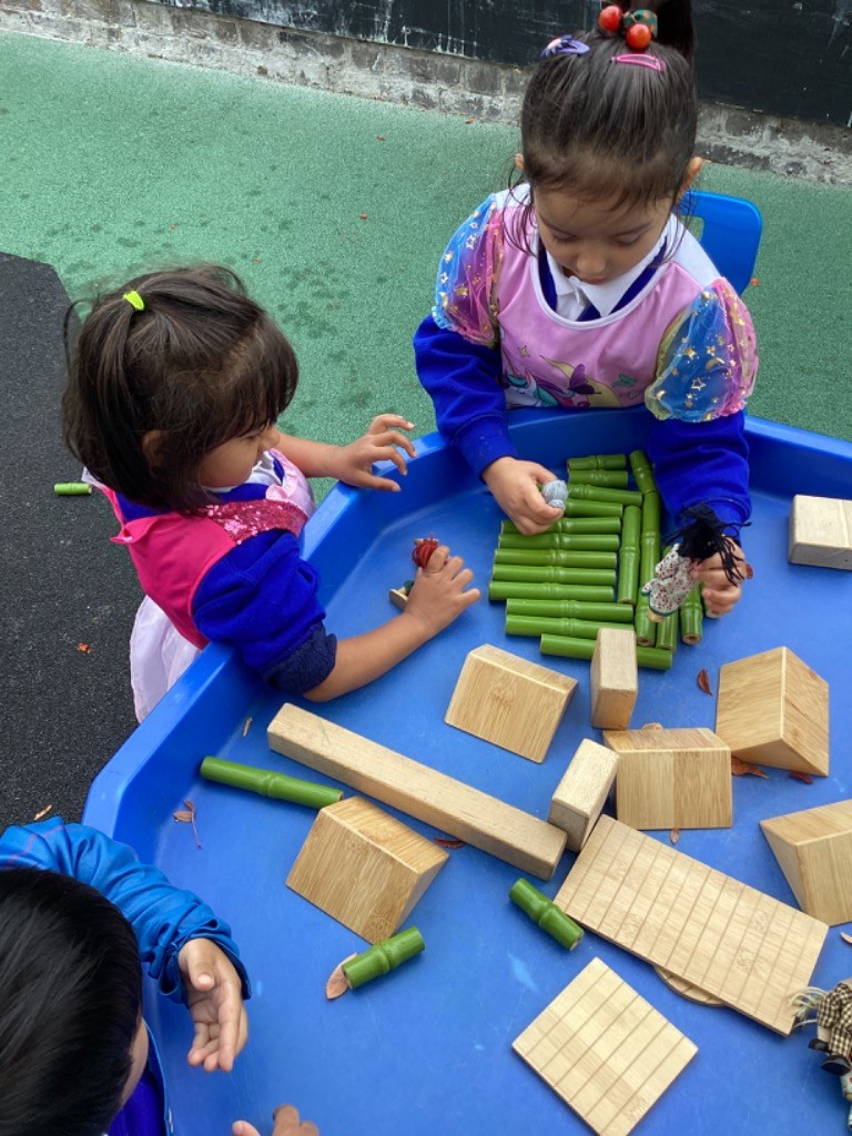 Two young girls are seated at a blue tray table, engaged in construction play with different materials. One child is building with green bamboo-like tubes and small wooden blocks, while the other is reaching for something on the table.