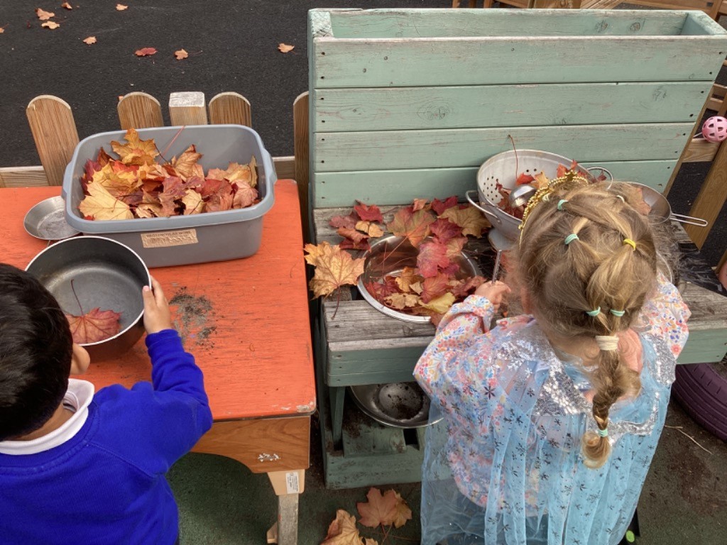 Two children are engaged in outdoor kitchen or imaginative play using natural materials. They are interacting with autumn leaves placed in bowls, a colander, and a plastic tub on a wooden play unit, possibly pretending to cook or make a potion.
