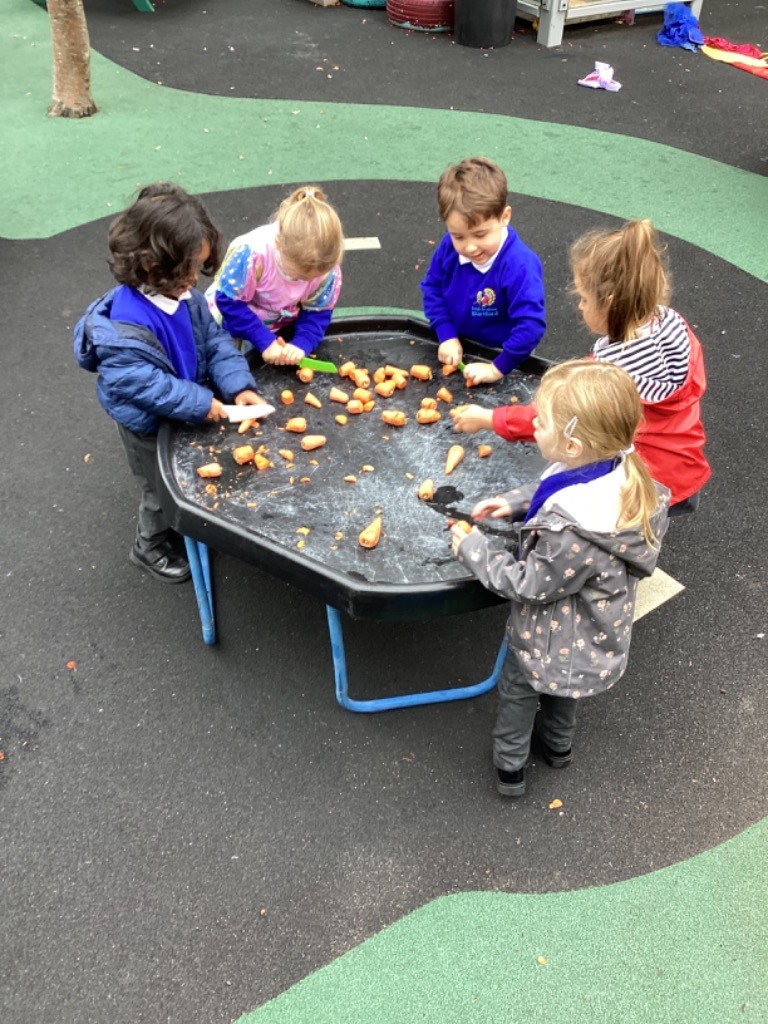 A group of five young children, dressed in a mix of school uniform and outdoor coats, are gathered around a black, hexagonal play tray (tuff tray) outdoors. The tray is scattered with orange pieces (possibly carrot pieces) and a bit of liquid or residue, suggesting a sensory or water play activity.