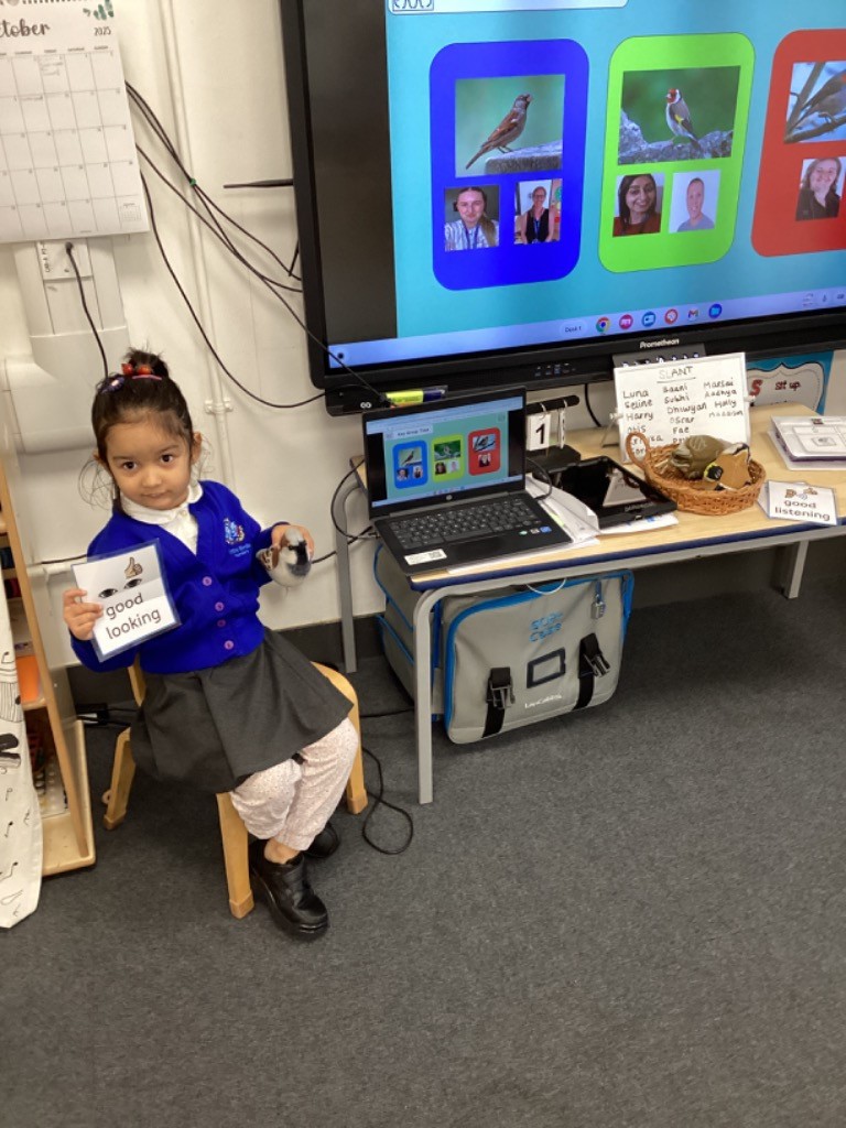 A young girl is sitting on a chair and holding up a card that says "good looking." The classroom setup, including the interactive screen displaying birds and the "Key Group Time" slide, is visible in the background.