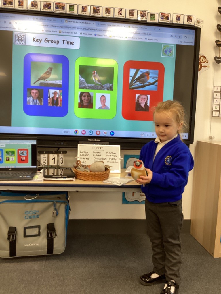 A young girl is standing in the classroom and holding a soft toy Goldfinch. The large interactive screen behind her shows the "Key Group Time" display, which features three different birds (a Sparrow, a Goldfinch, and a Robin) with pictures of staff members underneath, suggesting the children are divided into groups named after the birds.