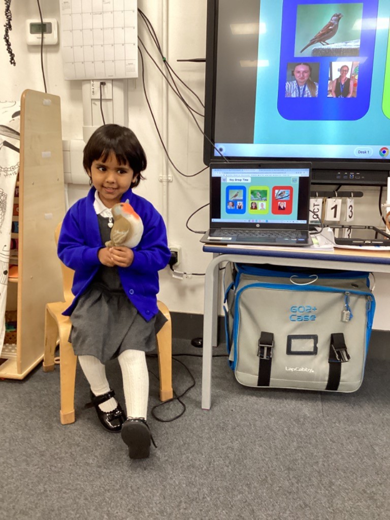 A young girl, wearing a school uniform, is sitting on a chair and holding a soft toy robin. She is smiling at the camera. In the background, there is a large interactive screen showing a "Key Group Time" slide with an image of a bird (likely the robin) and a laptop displaying the same slide.