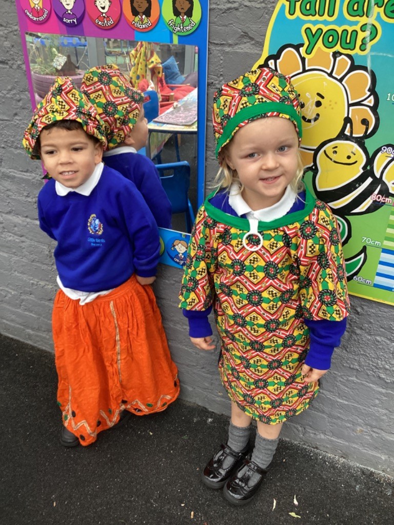 Two young children are standing outdoors, dressed in colorful, patterned clothing (over their school uniform) and matching hats, possibly for a celebration or dress-up day, and posing for the picture.