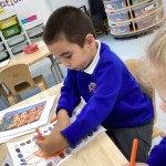 A young boy is seated at a table, concentrating on a coloring activity. He is coloring a printed sheet with an orange marker, and there's another image printout next to his work.