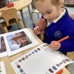 A young girl is seated at a table, drawing or writing with an orange marker. She is holding a small doll close to her, and there are other printed pictures on the table, possibly used as inspiration or part of a lesson.