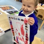 A young child is happily holding up a finished drawing or a picture they have been coloring with red and purple markers, smiling broadly at the camera.