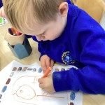 A young child is seated at a table, focused intently on coloring a picture with an orange marker or crayon. The picture looks like an outline of a bird or person. There's a container of colorful drawing tools next to the
