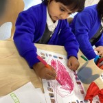 A young girl is focused on coloring a large shape (perhaps a bird or a leaf) on a piece of paper, using a pink or red marker to fill in the space with scribbled lines.