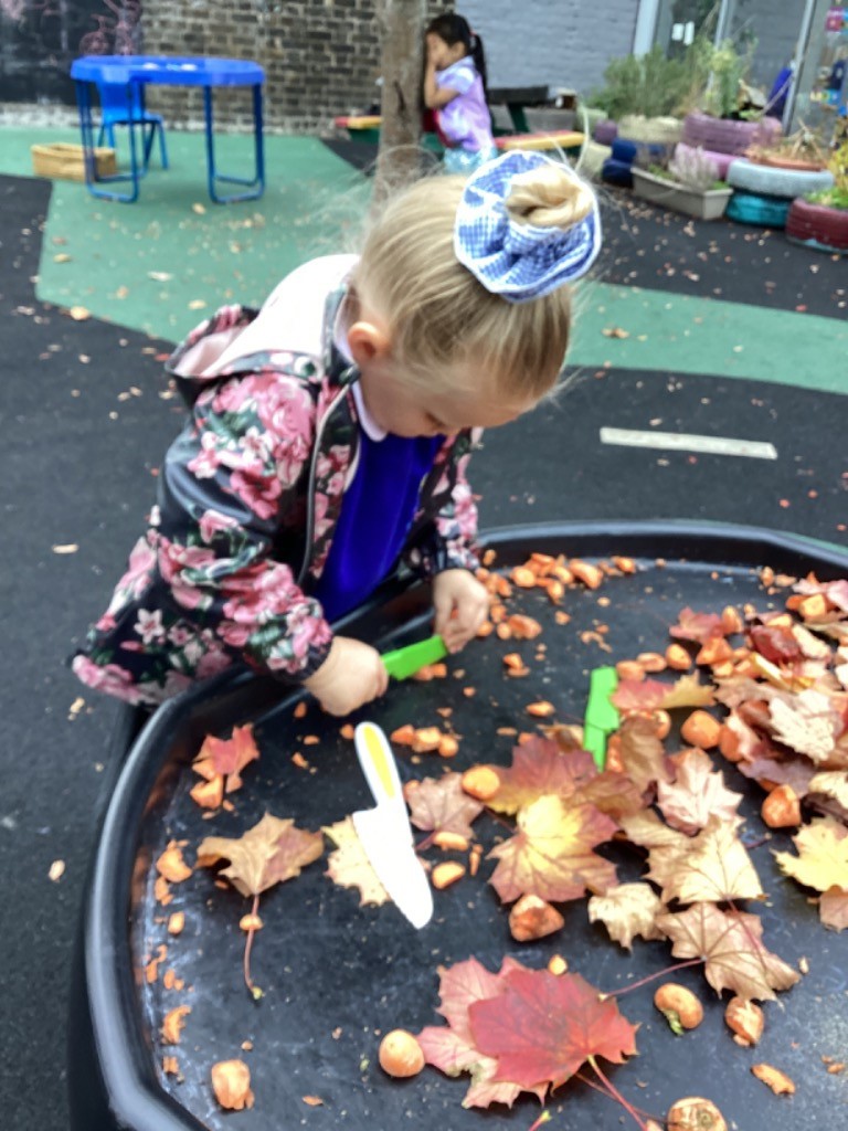 A young girl with her hair tied up is focused on an outdoor play activity. She's interacting with the leaves and small orange objects in the black tray, using a small white tool, possibly a toy knife or trowel. She is wearing a dark, floral print rain jacket.