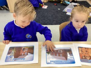 Two children are seated at a low table, looking at large flashcards or printouts featuring pictures of different house types, labeled as "detached" and "semi-detached." This suggests a lesson on housing and the built environment.