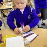 A child is using a glue stick to secure a white piece of paper to their craft stick construction, incorporating different materials into their building project.