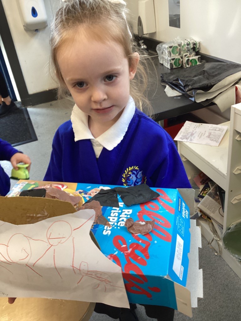 A child is holding up a creative construction project made from a cardboard box (looks like a Rice Krispies cereal box) that has been decorated with paper and brown clay or paint.