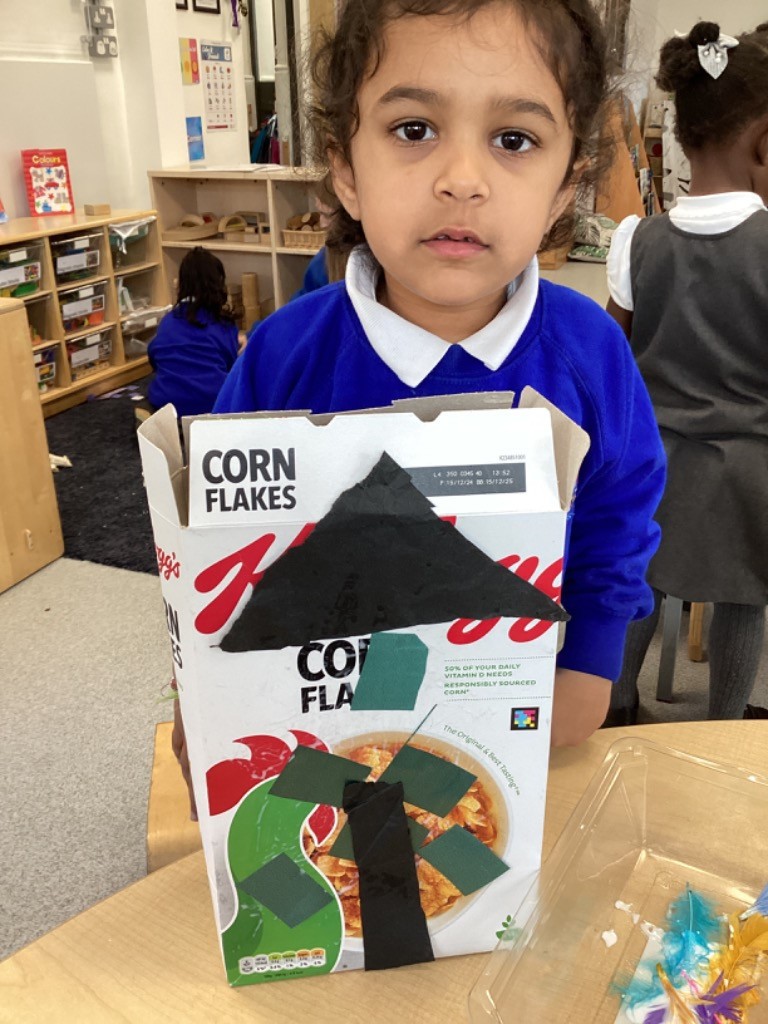 A child is holding a Corn Flakes cereal box that has been transformed into an art piece, decorated with black and green paper cutouts to resemble a roof and a tree.
