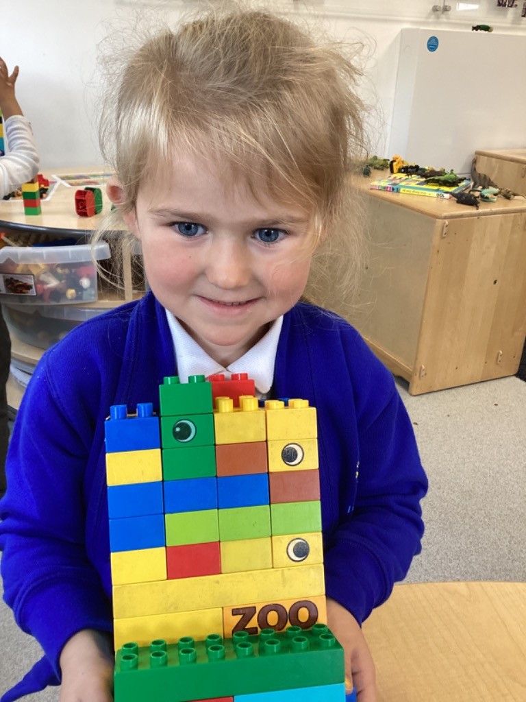 A child is happily holding a tall, colorful tower built out of large construction blocks (Duplo-style), with some bricks featuring animal eyes and the word "ZOO."