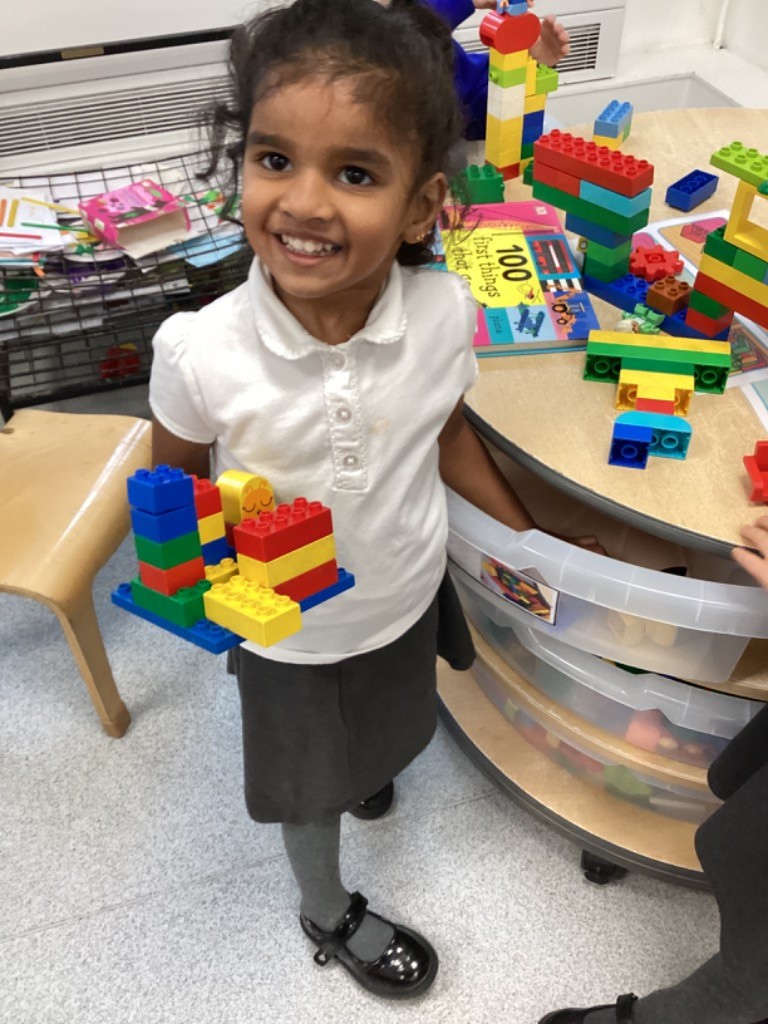 A child is smiling while holding a small, colorful structure they have built using large construction blocks. Other blocks and building materials are visible on the table behind her.