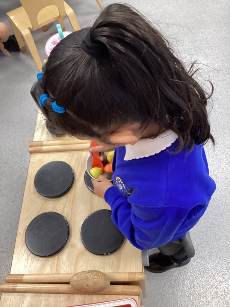 A child is focused on playing in a kitchen area, standing over a wooden play stove and handling a collection of toy fruit/food.