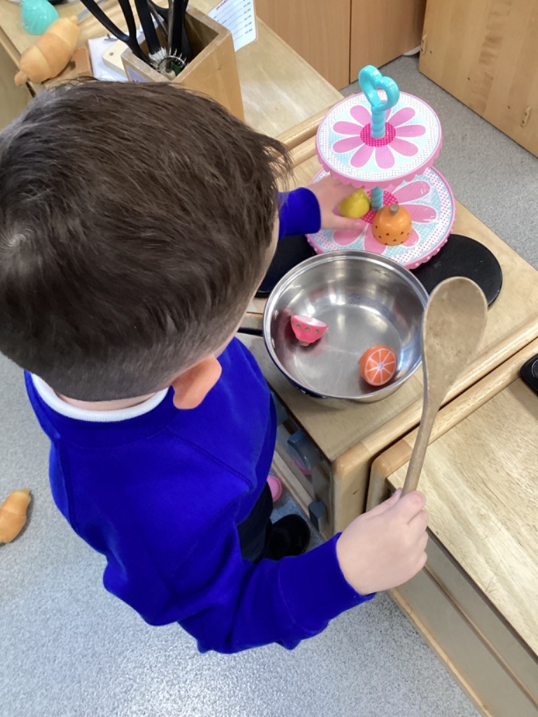 A child is using a large wooden spoon to "cook" in a small metal pot and is arranging toy fruit on a colorful tiered cake stand in the play kitchen area.