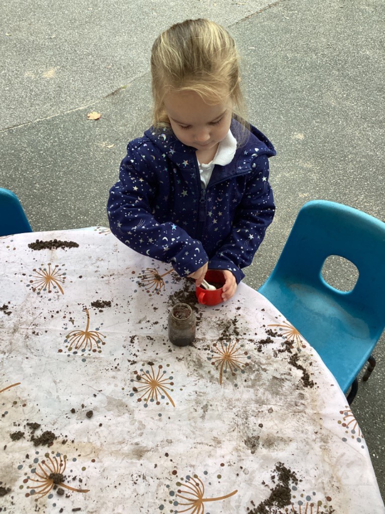 A child, wearing a coat, is engaged in sensory or outdoor play outside. They are sitting at a table covered in a mat and are using a spoon to scoop dirt or mud into a small red pot.
